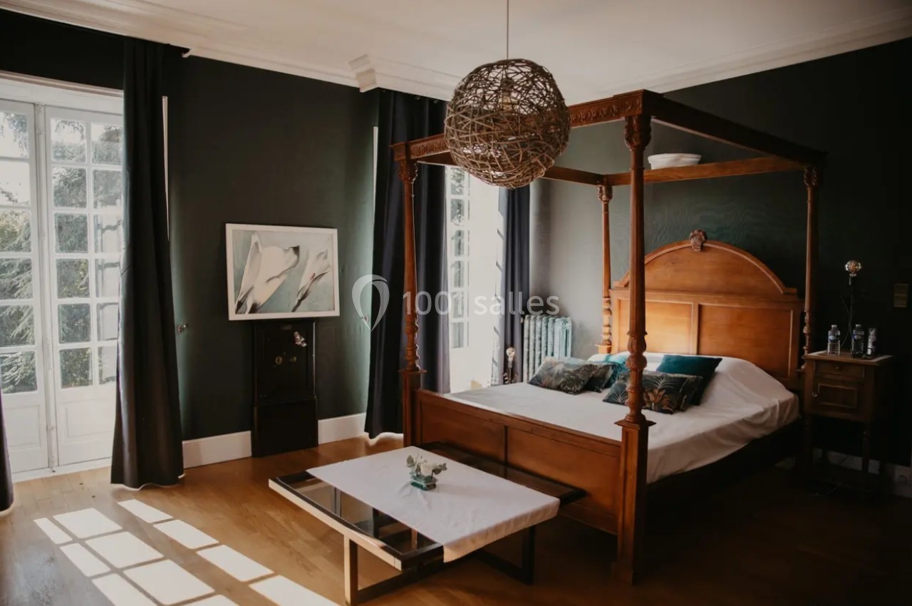 Chambre avec lit à baldaquin en bois, table basse, grandes fenêtres et murs sombres éclairés par la lumière naturelle.