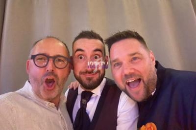 Trois hommes souriants posent ensemble devant un rideau beige, dans une ambiance conviviale.