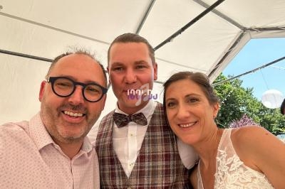 Un homme prend un selfie avec un couple souriant sous une tente blanche lors d'un événement en plein air.