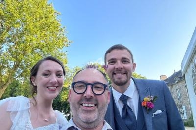 Un homme prend un selfie avec un couple souriant en tenue de mariage, dans un jardin par une journée ensoleillée.