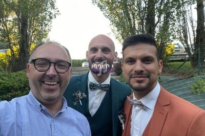 Trois hommes souriants en tenue formelle posent à l'extérieur devant des arbres et une clôture.