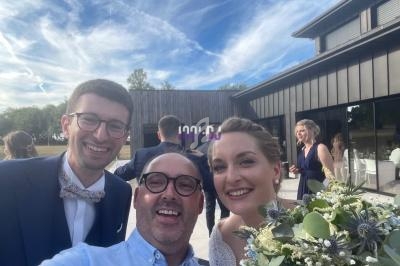 Un homme souriant prend une photo avec un couple en tenue de mariage devant un bâtiment moderne et un ciel dégagé.