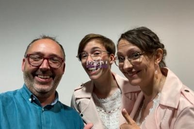 Trois personnes souriantes posant devant un mur clair, deux portant des vestes roses et une une chemise bleue.