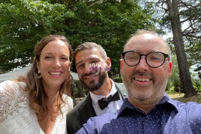 Un homme prend un selfie avec un couple souriant en tenue de mariage dans un jardin arboré.