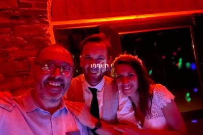 Un homme prend un selfie avec un couple souriant, dont la femme porte une robe blanche, dans une ambiance lumineuse rouge.