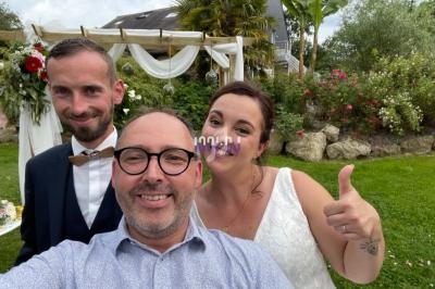 Un couple en tenue de mariage pose avec un homme souriant dans un jardin décoré de fleurs et de voilages.