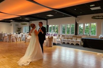 Un couple danse dans une grande salle lumineuse décorée pour une réception, avec des chaises alignées et des tables dressées.