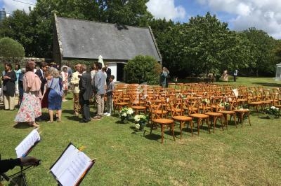 Chaises en bois disposées en extérieur pour une cérémonie, avec des invités rassemblés près d'un petit bâtiment.
