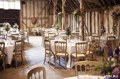 Salle de réception rustique avec tables rondes dressées, chaises dorées et décorations florales.