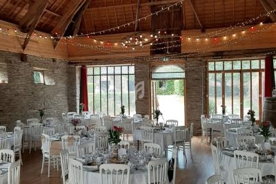 Salle de réception en pierre et bois, décorée avec des guirlandes lumineuses, tables rondes dressées avec nappes blanches.