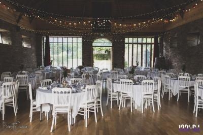 Salle de réception décorée avec des tables rondes dressées, chaises blanches et guirlandes lumineuses suspendues.