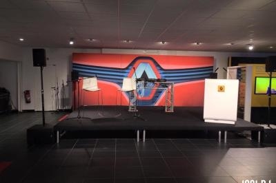 Scène avec pupitre, tabourets et équipement audio, devant un décor rouge et bleu avec un logo géométrique.