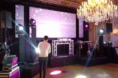 Un homme debout face à une scène illuminée avec écran géant, projecteurs et lustre dans une salle élégante.