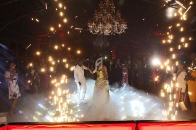 Un couple danse au centre d'une piste entourée d'effets pyrotechniques et de spectateurs dans une ambiance festive.