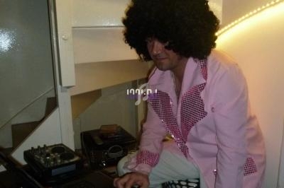 Homme en chemise rose et perruque afro utilisant un ordinateur portable près d'un escalier intérieur.