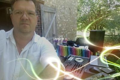 Homme en chemise blanche devant une table de mixage avec ordinateur, enceintes et décor extérieur verdoyant.