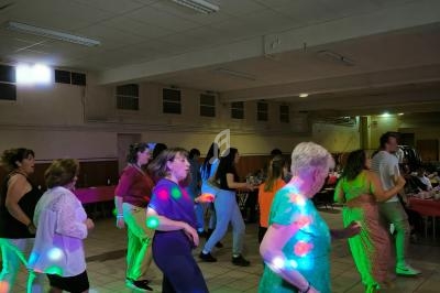 Des personnes dansent dans une salle éclairée par des spots colorés, avec des tables et des chaises en arrière-plan.