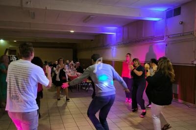 Des personnes dansent dans une salle éclairée par des lumières colorées, avec des tables et des convives en arrière-plan.