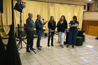 Un homme parle au micro devant quatre personnes sur une scène avec rideaux jaunes et équipement audio en arrière-plan.
