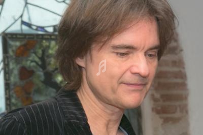 Homme aux cheveux mi-longs bruns, vêtu d'une veste rayée, devant un fond avec vitrail et mur en briques.