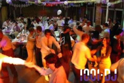 Des personnes dansent dans une salle décorée pour une fête, avec des tables dressées et des invités assis en arrière-plan.