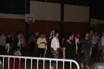 Des personnes dansent en ligne dans une salle de sport éclairée, avec des paniers de basket visibles en arrière-plan.