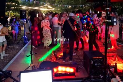 Groupe de personnes dansant en soirée en plein air avec éclairages colorés et un musicien jouant sur scène.