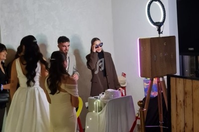 Des invités, dont une mariée en robe blanche, utilisent un photobooth dans une salle décorée avec des ballons et un…
