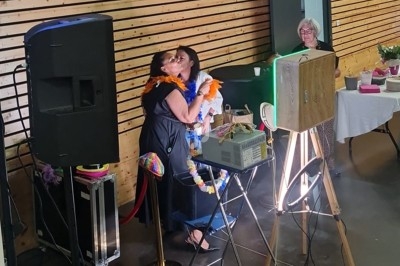 Deux personnes s'embrassent dans une salle avec des équipements audio et vidéo, sous le regard d'une femme en arrière-plan.