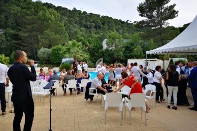 Groupe de personnes réunies en extérieur près d'une piscine, avec un chanteur et un micro au premier plan.