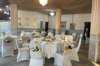 Salle élégante décorée pour un événement, avec tables rondes dressées, chaises blanches et éclairage chaleureux.