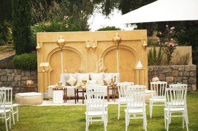 Décoration extérieure avec canapé, lanternes dorées et chaises blanches disposées devant un mur sculpté.