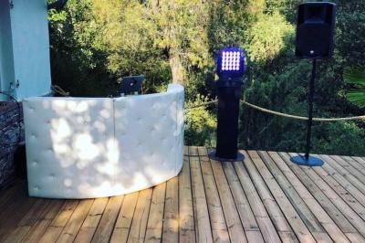 Poste de DJ avec table blanche, éclairage et enceintes installé sur une terrasse en bois entourée de végétation.