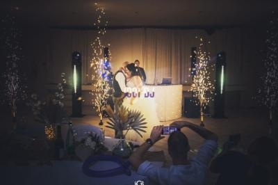 Un DJ anime une soirée festive avec des effets lumineux et des étincelles, devant des invités dans une salle décorée.