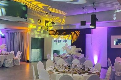 Salle de réception décorée avec des tables rondes dressées, des chaises blanches et un éclairage coloré.