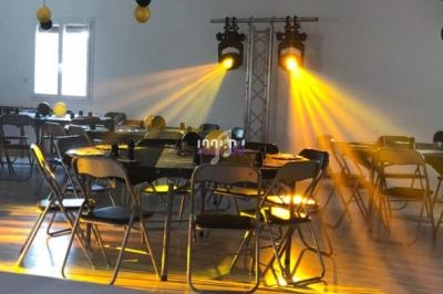 Salle décorée avec des ballons noirs et jaunes, tables et chaises disposées, éclairage jaune vif projeté au sol.