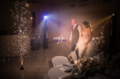 Un couple de mariés regarde un spectacle lumineux avec des étincelles dans une salle de réception décorée.