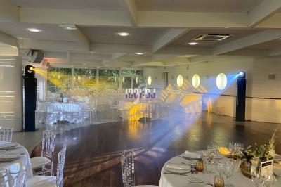 Salle de réception lumineuse avec tables dressées, chaises transparentes et éclairage coloré projeté sur le sol.