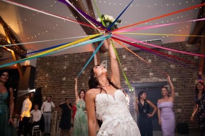 Une mariée lève un bouquet de fleurs au centre de rubans colorés tenus par des invités lors d'une célébration.