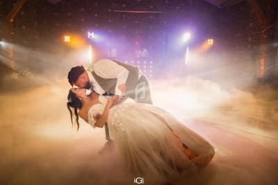 Un couple de mariés danse dans une salle éclairée, entouré de fumée et de lumières colorées.