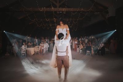 Un couple danse lors d'un mariage, l'homme soulève la femme sous des lumières et une ambiance brumeuse.