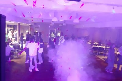 Salle de réception animée avec des invités dansant sous des lumières colorées et de la fumée au sol.