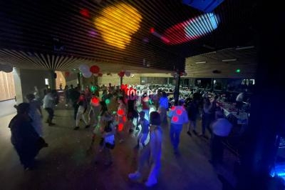 Des personnes dansent dans une salle sombre éclairée par des lumières colorées, avec des tables en arrière-plan.