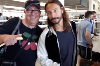 Deux hommes posent ensemble dans un aéroport, l'un tenant un passeport, des passagers et bagages en arrière-plan.