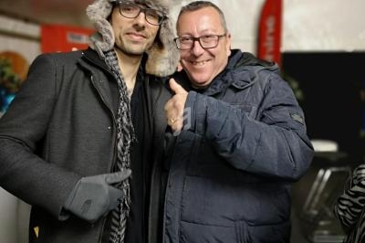 Deux hommes souriants posent ensemble sous une tente, l'un portant un manteau et un chapeau en fourrure.