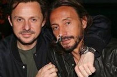 Deux hommes posant côte à côte, l'un avec un bras autour de l'épaule de l'autre, dans un cadre intérieur sombre.