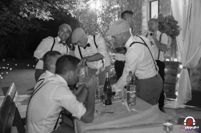 Des hommes en tenue rétro discutent et rient autour d'une table lors d'un événement en extérieur, éclairé par des lumières.