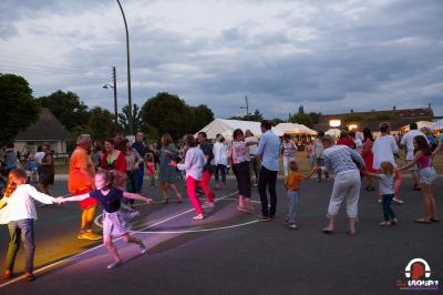 Des personnes dansent en plein air sur une place, entourées de tentes blanches et d'arbres, en soirée.