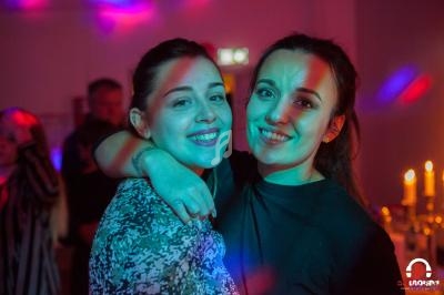 Deux femmes souriantes posent ensemble dans une ambiance festive avec éclairages colorés et bougies en arrière-plan.