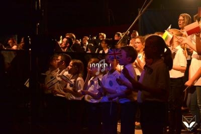 Des enfants et adultes chantent en chœur sur scène, certains tenant des drapeaux de différents pays.